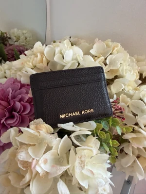 korthållare från Michael Kors - Stilren mörkbrun korthållare från Michael Kors i präglat skinn med guldfärgad logga framtill. Praktisk och smidig modell med flera kortfack, perfekt för dig som vill ha koll på korten utan att bära en stor plånbok.