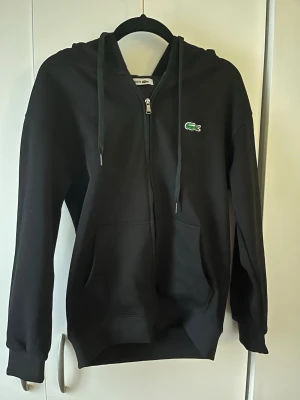 Svart zip-hoodie från Lacoste - Svart hoodie med dragkedja från Lacoste, klassisk krokodillogga på bröstet och stora fickor framtill. Mjuk bomullskänsla och justerbar huva med snören. Perfekt för en chill och stilren look. Pris lapp finns helt ny tröja 