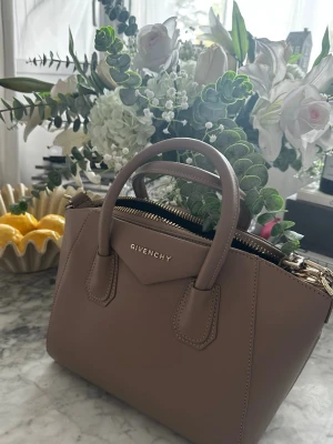 Beige handväska vintage - Snygg beige handväska i slätt läderimitation med guldfärgade metalldetaljer och två topphandtag. Väskan har en strukturerad form och dragkedja upptill. Perfekt för dig som gillar lyxiga accessoarer med stilren design. Vintage NAKD, HM, Zara