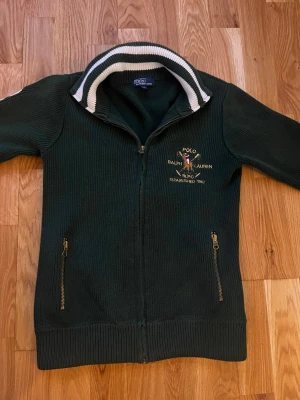 Grön stickad ziptröja Polo Ralph Lauren - Mörkgrön stickad ziptröja från Polo Ralph Lauren med broderad logga och text på bröstet. Tröjan har ståkrage med vita ränder, två fickor med dragkedja och ribbade muddar. Perfekt för en sportig och stilren look.