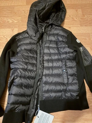 Svart pufferjacka från Moncler - Snygg svart pufferjacka från Moncler med stickade ärmar och huva. Jackan har dragkedja framtill, två fickor med knappar och en extra ficka på ärmen. Klassisk Moncler-logga på armen och quiltad design på fram- och baksidan. Perfekt för kyliga dagar.