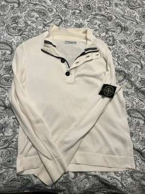 Vit långärmad tröja från Stone Island - Vit långärmad tröja från Stone Island med svart logotyp-patch på ärmen. Tröjan har hög krage med flera svarta knappar och ribbade muddar. Snygga svarta detaljer på kragen ger en sportig känsla. Perfekt för dig som gillar streetwear.