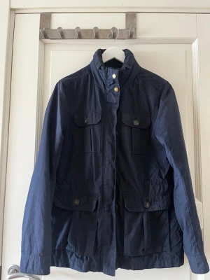 Mörkblå vårjacka med fickor - Stilren mörkblå vindjacka med hög krage, luva, dragkedja och knappar framtill. Fyra stora fickor med lock och knappstängning ger en cool utility-look. Perfekt för blåsiga dagar och enkel att matcha med olika outfits.