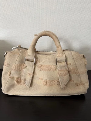 Beige distressed handväska - Cool beige handväska i canvas med slitna detaljer och silverfärgade metalldelar. Väskan har två rejäla handtag och dragkedja upptill. Den unika distressed looken ger väskan en trendig och avslappnad vibe. Perfekt storlek för att få plats allt man behöver men inte för stor.