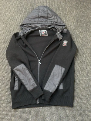 Svart Cardigan  - Svart hoodie med dragkedja. Har quiltad huva och detaljer i blankt tyg på ärmar och fickor. Märkets logga på bröstet och insidan av kragen. Perfekt för en chill och stilren look. Perfekt inför sommaren med en luva som man enkelt kan ta av. Storlek M. Mycket fint skick förutom ett litet hål i ena armbågen.