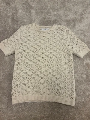 Beige stickad topp från Mango - Snygg beige stickad topp från Mango med korta ärmar och ett detaljerat hålmönster över hela plagget. Rund halsringning och ribbade kanter ger en clean look. Perfekt till jeans eller kjol för en trendig stil.