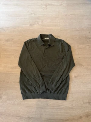 Mörkgrön pikétröja med lång ärm - Stilren mörkgrön pikétröja med lång ärm och klassisk krage. Tröjan har knäppning framtill och ribbade muddar vid ärmslut och nederkant. Perfekt för en avslappnad men snygg look.