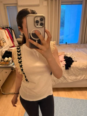 Vit ärmlös topp med hjärtdetaljer - Supersöt vit ärmlös topp från Love Lolita med svarta hjärtmönster och beige fransar längs axlarna. Perfekt för dig som vill ha något unikt och lekfullt i garderoben. Toppens material känns mjukt och bekvämt mot huden.