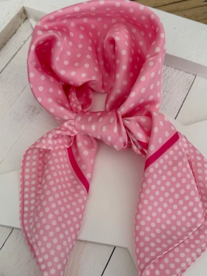 Pink Soufflé – Polka Scarf 70x70 - Mjuk rosa scarf med fina vita prickar och elegant ceriserosa kant – en feminin klassiker med lekfull och romantisk känsla. Den satinlika finishen ger ett mjukt lyster och ett följsamt fall som gör scarfen lätt att bära både till vardag och sommarens ljusa kvällar.  Den puderrosa tonen tillsammans med de små prickarna ger en tidlös vintagestil med modern elegans – lika fin knuten runt halsen som i håret eller på väskan.  • 70 x 70 cm • Rosa med vita prickar och ceriserosa detaljer • Satin finish 