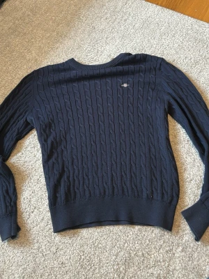 Mörkblå kabelstickad tröja Gant - Kabelstickad mörkblå tröja från Gant med rund halsringning och broderad logga på bröstet. Tröjan har ribbade muddar vid ärmslut och nederkant, och är tillverkad i ett mjukt stickat material som ger en klassisk och stilren look.
