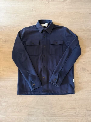 Mörkblå overshirt från Selected - Stilren mörkblå overshirt från Selected med klassisk krage, två bröstfickor med lock och knappar framtill. Tillverkad i ett kraftigare tyg som ger en snygg och strukturerad look. Perfekt att bära öppen eller stängd över en t-shirt.