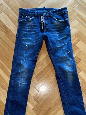Mörkblå Dsquared2 Jeans - Snygga mörkblå jeans från Dsquared2 med slitningar och färgstänk för en cool, trendig look. Klassisk femficksmodell med dragkedja och röd Dsquared2-tag vid gylfen. Perfekta för dig som gillar streetstyle och detaljer.