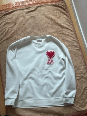 Vit sweatshirt från Ami Paris - Vit sweatshirt från Ami Paris .Tröjan är i väldigt bra skick Ja köpte den i februari 2026 alltså ja har inte haft den länge riktigt fin tröja i stl L.Den kommer också med sitt orginal packet och påse