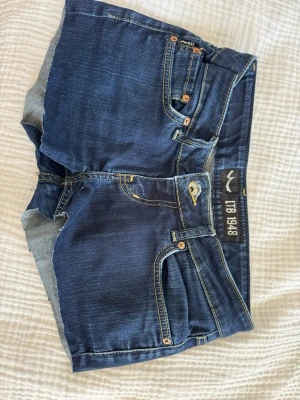 Mörkblå jeansshorts från LTB - Snygga mörkblå jeansshorts från LTB med klassiska fem fickor, gul kontrastsöm och broderad detalj på bakfickorna. Shortsen har låg midja och är gjorda i mjuk bomullsdenim med en bekväm passform. Perfekta för varma dagar!