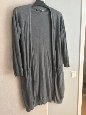 Grå lång cardigan med fickor - Mysig och enkel grå lång cardigan med två fickor framtill. Öppen modell utan knappar, perfekt att slänga på sig över en t-shirt eller topp. Långärmad och i mjukt stickat material som känns skönt mot huden.