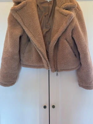 Beige teddyjacka från NA-KD - Säljer en supermjuk och fluffig beige teddyjacka från NA-KD. Jackan har dragkedja framtill, två fickor med dragkedja och en bred krage. Perfekt för att hålla sig varm och snygg under kyliga dagar. Materialet är fuskpäls som ger en mysig känsla.
