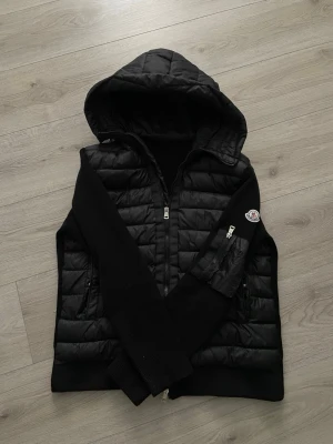 Svart Monclear cardigan - Svart cardiganjacka  med vadderad front och huva. Ärmar och rygg i stickat material, medan framsidan är quiltad och glansig. Jackan har dragkedja framtill och en . Perfekt för lager-på-lager-stil. NFC FUNKAR