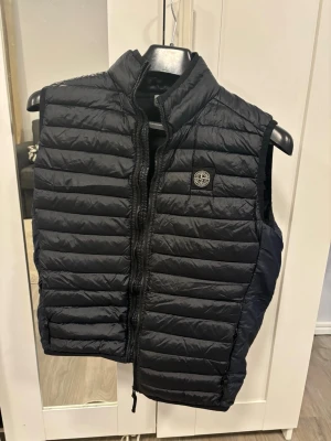 Svart dunväst från Stone Island - Snygg svart dunväst från Stone Island med quiltad design och dragkedja framtill. Västen har en hög krage och den ikoniska Stone Island-loggan på bröstet. Perfekt att slänga över en hoodie eller tröja för en cool och avslappnad look.