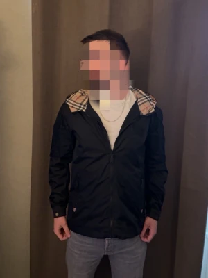 Svart vindjacka med rutigt foder - Snygg svart vindjacka med dragkedja och huva. Insidan av huvan har ett klassiskt beige-rutigt mönster. Jackan har två fickor framtill och är tillverkad i ett lätt, vattenavvisande material. Perfekt för blåsiga dagar och enkel att matcha med olika outfits.