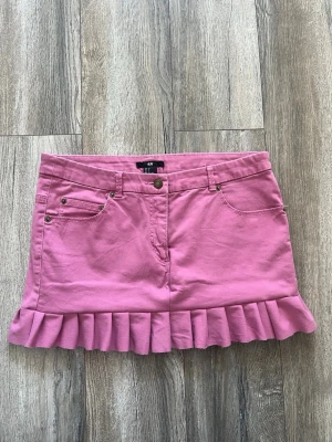 Rosa volangkjol från H&M - Superfin rosa kortkjol från H&M med volangkant nertill. Kjolen har klassisk jeansmodell med fickor fram och bak samt hällor för skärp. Perfekt för dig som vill ha en söt och trendig look.
