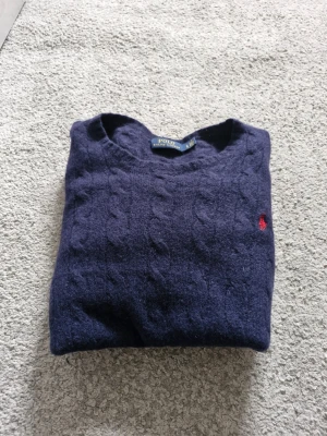 Polo Ralph Lauren tröja - Kabelstickad mörkblå tröja från Polo Ralph Lauren med klassisk rund halsringning och röd broderad logga på bröstet. Tröjan har långa ärmar och ribbade muddar vid ärmslut och nederkant. Perfekt för en stilren och avslappnad look. Har bara ett litet hål på armen. Syns knappt. Annars bra skick