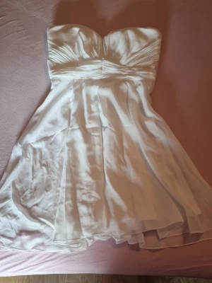 Vit axelbandslös kortklänning - Snygg vit axelbandslös klänning med draperad överdel och lätt utställd kjol. Klänningen har en romantisk känsla med subtila paljett-detaljer vid midjan och är tillverkad i ett tunt, svepande material som ger ett elegant fall. Helt ny med prislapp