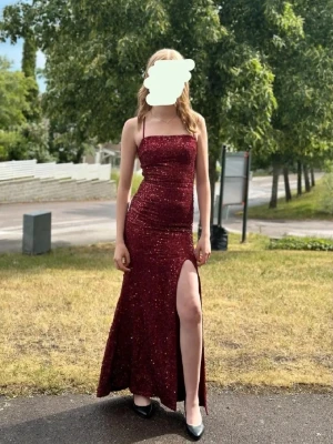 Vinröd glittrig långklänning med slits - Vinröd långklänning med glittrigt tyg, smala axelband och snörning i ryggen. Klänningen har en figurnära passform och en hög slits framtill som ger en elegant look. Perfekt för bal eller festliga tillfällen. Nypris 460kr, använd 1 gång. 