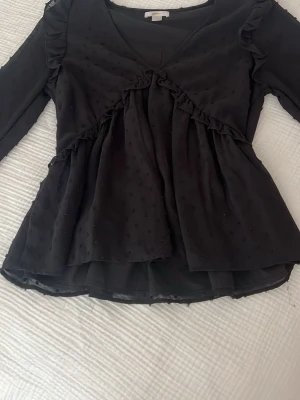 Svart blus med volang från Gina Tricot - Supersöt svart blus från Gina Tricot med volangdetaljer och v-ringning. Lätt transparent tyg med diskret prickstruktur och långa ärmar. Perfekt att styla med jeans eller kjol för en trendig look.