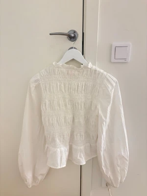 Vit blus med smock och puffärmar - Superfin vit blus med smock över bröstet, puffiga långärmar och volang nertill. Blusen har knappar framtill. Perfekt till vardags och till fest!