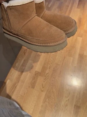 Rosemunde teddy boots  - Mysiga bruna tofflor i mocka med mjukt fårskinnsfoder och kraftig sula. Klassisk UGG-inspirerad design med rund tå och låg skaft. Perfekta för att hålla fötterna varma hemma under kalla dagar.