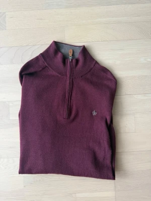 Vinröd halvzip-tröja från Morris - Snygg vinröd stickad tröja från Morris med halv dragkedja och ståkrage. Tröjan har en liten broderad logga på bröstet och är fodrad i grått på insidan av kragen. Perfekt för lager-på-lager och stilrena outfits.