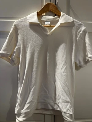 Vit ribbad polotopp med krage - Vit ribbad polotopp med kort ärm och klassisk krage. Toppen har en v-ringad öppning framtill och är tillverkad i ett mjukt, bekvämt material. Perfekt för en stilren och avslappnad look.