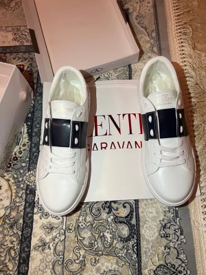 Valentino vita sneakers med svart band - Snygga vita sneakers från Valentino med ett brett svart band över vristen och silverfärgade nitar. Skorna har klassisk låg profil, vita skosnören och är tillverkade i slätt läder. Perfekta för dig som vill ha en clean men ändå edgy look.
