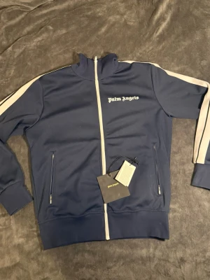 Blå ziptröja från Palm Angels - Mörkblå ziptröja från Palm Angels med vita ränder längs ärmarna och vit dragkedja. Tröjan har hög krage, två fickor med dragkedja och logga på bröstet. Perfekt för en sportig och trendig look.
