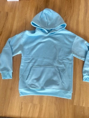 Ljusblå hoodie med magficka - Snygg ljusblå hoodie med klassisk huva och stor magficka framtill. Tillverkad i mjukt material som känns skönt mot huden. Perfekt för dig som gillar en enkel och clean stil. Långa ärmar med ribbade muddar och avslappnad passform.