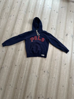 Marinblå hoodie från Polo Ralph Lauren - Snygg marinblå hoodie från Polo Ralph Lauren. Klassisk känguruficka och vit dragsko i huvan. Perfekt för en avslappnad och sportig stil. Mjuk bomullskvalitet och bekväm passform. Mycket fint skick, använd fåtal gånger. Storlek S