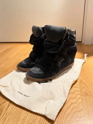 Svarta Isabel Marant wedge sneakers - Snygga svarta sneakers från Isabel Marant med inbyggd kilklack. Skorna har en cool design med kardborreband, mockadetaljer och gummisula. Perfekta för dig som vill ha både stil och höjd i ett par sneakers. Ena sulan är LITE lös och en annan sula då den andra gick sönder 
