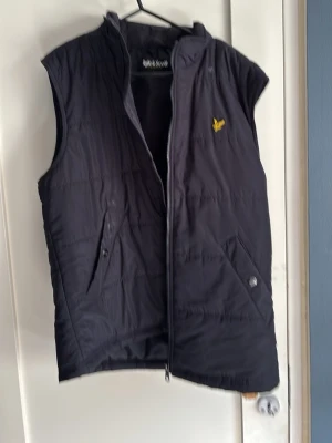 Svart dunväst från Lyle & Scott - Svart dunväst från Lyle & Scott med dragkedja framtill och två snedställda fickor. Klassisk gul logga på bröstet och hög krage. Perfekt för lager-på-lager och streetwear-stil. Materialet är lätt och quiltat för extra värme. Funkar som m