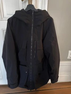 Svart vindjacka Peak Performance - Snygg svart vindjacka från Peak Performance med huva och dragkedja framtill. Jackan har flera praktiska fickor, både med dragkedja och lock, samt justerbara ärmslut. Perfekt för blåsiga dagar och enkel att matcha med olika outfits. Nypris: 4000kr