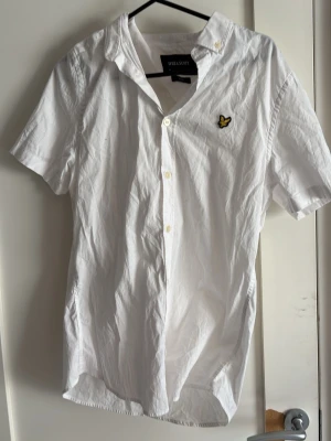 Vit kortärmad skjorta Lyle & Scott - Vit skjorta från Lyle & Scott i slim fit-modell med korta ärmar och klassisk krage. Skjortan har knappar framtill och den ikoniska gula loggan broderad på bröstet. Perfekt för en clean och stilren look.