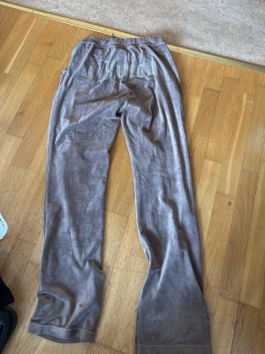 Bruna Juicy Couture mjukisbyxor - Säljer ett par bruna mjukisbyxor från Juicy Couture i mjuk velour. Byxorna har raka ben, resår i midjan med dragsko och fickor fram. På baksidan står det 'Juicy' i glittriga stenar. Perfekta för en chill och avslappnad stil.