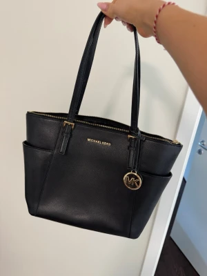 Svart Michael Kors handväska i läder - Snygg svart handväska från Michael Kors i strukturerat läder med guldfärgade detaljer och MK-hänge. Väskan har två långa axelband, dragkedja upptill och praktiska ytterfack på sidorna. Perfekt för dig som vill ha en stilren och rymlig väska.