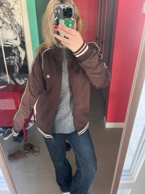 Brun track jacket från Dickies - Säljer en brun track jacket från Dickies med vita ränder längs ärmarna och muddar. Jackan har dragkedja framtill, ståkrage och ribbade muddar vid ärmslut och nederkant. (Dragkedjan har ingen dragare men funkar ändå) 