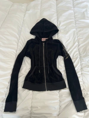 Svart velour hoodie Juicy Couture - Svart velour hoodie från Juicy Couture med dragkedja, huva och snörning. Stora guldfärgade broderier på ryggen och ärmen ger en lyxig vibe. Fickor framtill och ribbade muddar. Perfekt för en trendig och avslappnad look.