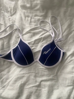 Blå bikiniöverdel från Panos Emporio - Snygg bikiniöverdel från Panos Emporio i marinblått med vita kanter och tunna vita linjer. Klassisk trekantsmodell med justerbara axelband och diskret logga på ena kupan. Perfekt för strandhäng och pooldagar.