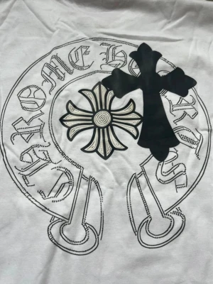 Vit Chrome Hearts t-shirt med tryck - Vit t-shirt från Chrome Hearts med svart och beige tryck framtill. Trycket består av ett stort kors och en blomliknande detalj, samt text i en cirkel. Klassisk passform och korta ärmar. Perfekt för dig som gillar streetwear och unika prints.