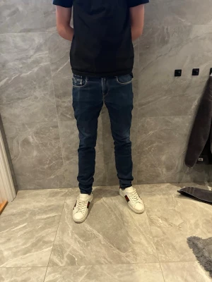 Mörkblå slim fit jeans replay  - Säljer ett par mörkblå slim fit jeans med vita kontrastsömmar och klassiska fem fickor. Jeansen har en modern passform som sitter tajt längs benen och är perfekta för en avslappnad men ändå stilren look.