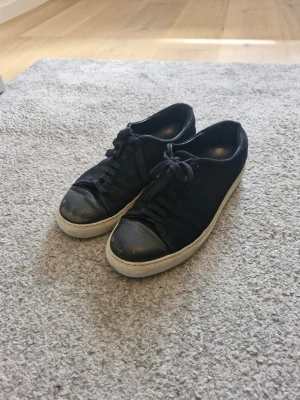 Axel Arigato skor - Stilrena svarta sneakers från Axel arigato med svart ovandel i tyg och detaljer i läder på tån. Skorna har vita sulor och klassisk låg profil med svarta skosnören. Perfekta för en clean och enkel look. Den är lite sönder vid sidan och därför är priset så. Hör av dig av frågor