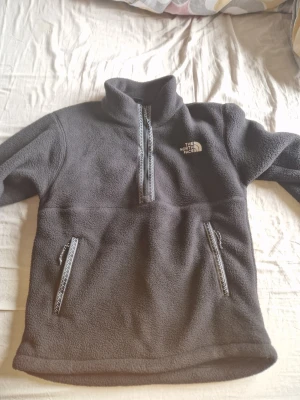 Grå fleece från The North Face - Mysig grå fleece med halv dragkedja från The North Face. Tröjan har två fickor med dragkedja och hög krage. Perfekt för lager-på-lager och outdoor-looks. Klassisk logga på bröstet och detaljerade dragkedjor.