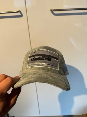 Grå velvet keps  - Säljer en grå velvet keps med ett coolt bilmotiv på framsidan. Kepsen har böjd skärm och mesh baktill för extra ventilation. Justerbar storlek med snapback-spänne. 
